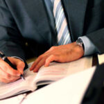 A person in a suit writing in a notebook with a pen.