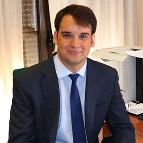 Man in a suit sitting at an office desk, smiling.