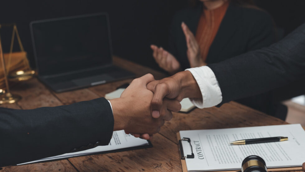 Two professionals shaking hands across a wooden table with a laptop and documents nearby.