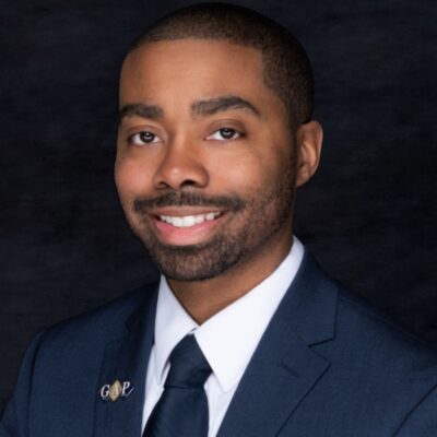 Professional headshot of a smiling man in a blue suit and tie against a dark background. Professional headshot of a smiling man in a blue suit and tie against a dark background.